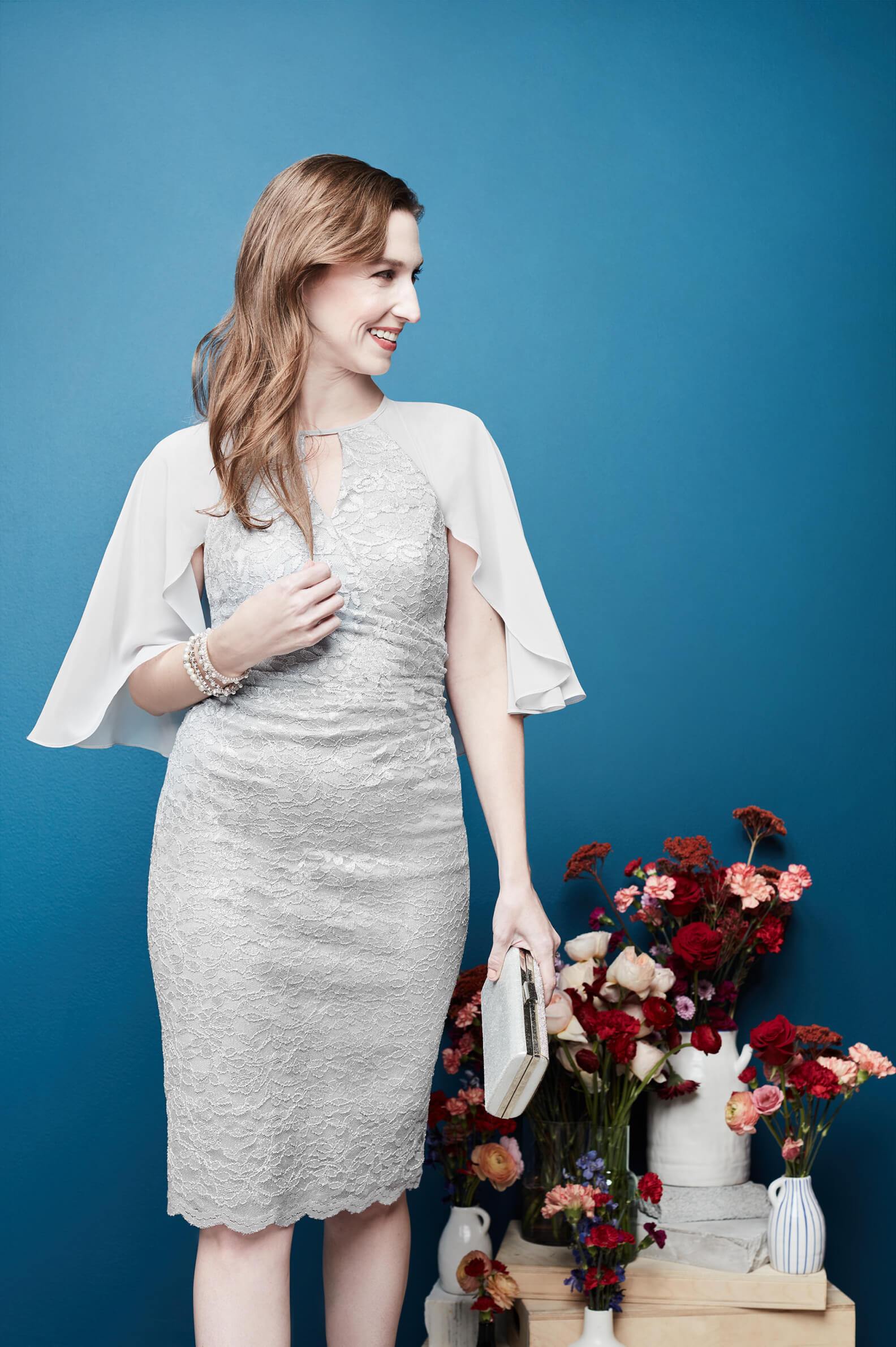 woman in a silver dress and capelet holding a clutch standing in front of a table with flowers in vases on it